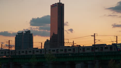 El-Tren-Subterráneo-De-Seúl-Viaja-Por-El-Puente-Ferroviario-Hangang-Al-Atardecer,-El-Edificio-63-Contra-El-Impresionante-Fondo-De-Cielo-Naranja-Degradado-Colorido