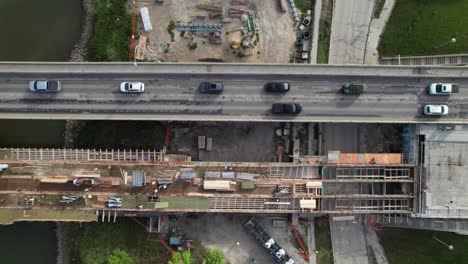 Overhead-of-bridge-construction-site,-side-by-side-with-active-highway,-4K-drone-shot