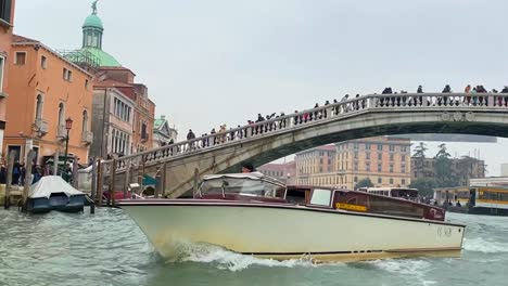 Pullback-under-crowded-venice-canal-bridge-as-ferry-cuts-across-bustling-waterway