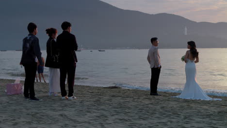 Fotógrafo-De-Bodas-Posa-A-Una-Hermosa-Pareja-Asiática-Al-Atardecer-Mientras-Las-Olas-Del-Océano-Rompen-En-La-Playa