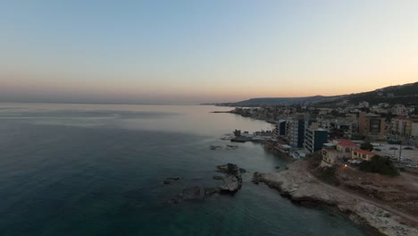 Drone-flight-along-coastline-of-Batroun-City-with-luxury-buildings-and-tranquil-mediterranen-sea-at-sunset-time,-Lebanon