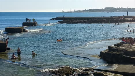 Piscinas-Naturales-De-Agua-De-Mar-Con-Jóvenes-Al-Fondo-Saltando-De-Las-Rocas-Al-Agua-Azul