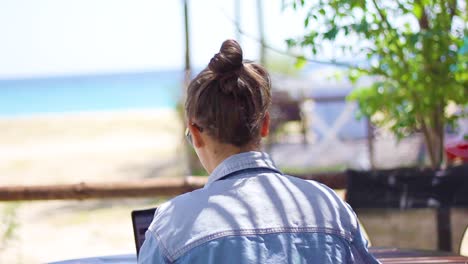Woman-working-on-laptop-seen-from-behind-at-beach-with-sea-view