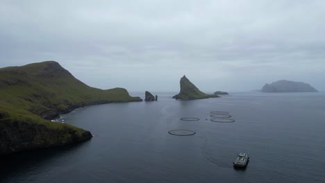 Salmon-aquaculture-farm-with-fishing-boat-front-Drangarnir-and-Tindholmur-in-Vagar,-Faroe-Islands