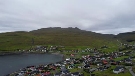 Sandavagur-coastal-village-at-base-of-rolling-mountain-hillsides,-aerial-aescend-in-Faroe-Islands