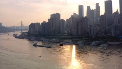 Golden-hour-sunset-light-cuts-through-skyscrapers-as-barge-and-tram-cross-sky-and-river,-Chongqing-city-china,-aerial