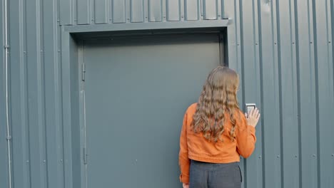 Brown-haired-woman-in-orange-jacket-walks-towards-urban-building,-pushes-key-code,-rear-view