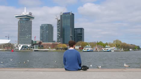 Professional-Photographer-Sitting-By-The-IJ-River-Next-To-Analog-Film-Camera-In-Amsterdam,-Netherlands