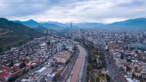 Aerial-hyperlapse-doll-out-with-business-center-in-the-center-and-cloudy-skyline-with-snowy-Andes-mountain-range,-high-vehicular-traffic