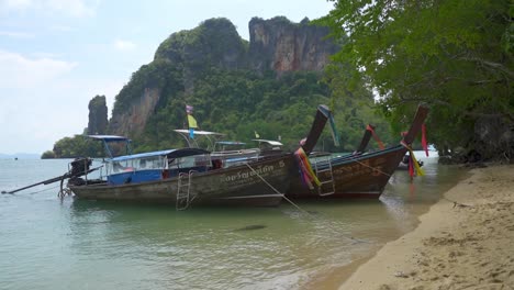 LongTail-Thailand-Boats-Anchored,-flags-waving,-with-tropical-island-cliffs-behind