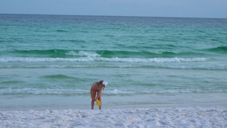 Little-girl-wearing-swimsuit-plays-with-water-and-sand-on-the-beach-with-shovel-in-the-hand