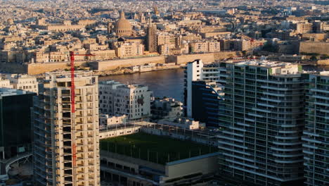 Aerial-tilt-shot-of-condos-and-a-soccer-field-with-the-sunset-Malta-skyline-background
