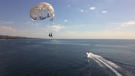 Luftaufnahme-Von-Parasailing-In-Marbella-Malaga,-Lustige-Wasseraktivität-An-Einem-Sonnigen-Tag,-Schnelles-Boot,-Das-Mit-Zwei-Personen-Einen-Mistral-Strand-Fallschirm-Zieht,-Blauer-Himmel-In-Spanien,-4K-Aufnahme