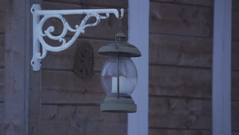 Retro-metal-lantern-with-a-candle-inside,-hanging-from-a-wooden-house-and-swinging-in-the-wind,-at-night
