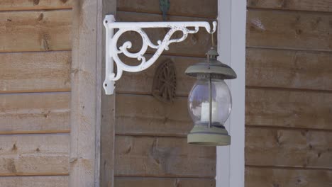 Detail-of-a-retro-metal-lantern-with-a-candle-inside,-hanging-from-a-wooden-house-and-swinging-in-the-wind