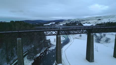 Aerial-Drone-Ascends-Captivating-a-Tilt-Down-Panorama-of-Road-and-Rail-Bridge-Spanning-the-Tranquil-River-Findhorn-in-Tomatin,-Near-Inverness,-Adorned-by-a-Blanket-of-Snow-Covered-Highland-Scenery