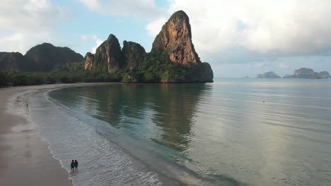 Railay-Beach-Thailand,-Two-people-walking-by-the-ocean-at-sunrise-with-limestone-cliffs--aerial