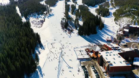 People-Skiing-and-Riding-Chairlifts-down-Ski-Slope-next-to-Parking-Lot-and-Pine-Tree-Forest-and-Snow