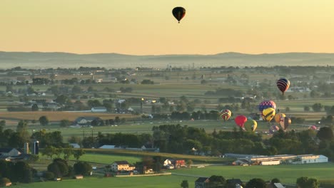 Aerial-wide-shot-showing-many-hot-air-balloons-starting-on-green-scenic-field-in-american-countryside-at-sunrise