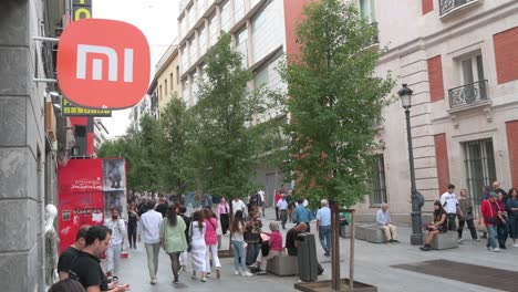 People-walk-past-the-Chinese-multinational-technology-and-electronics-brand-Xiaomi-flagship-store-and-its-logo-sign