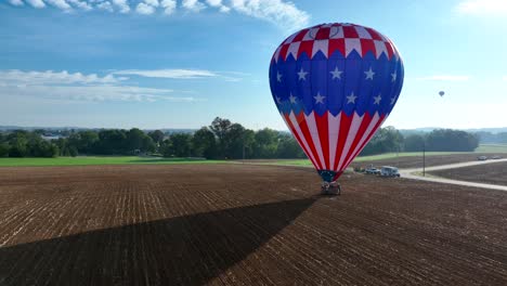 Aerial-approaching-shot-of-american-hot-air-balloon-landing-on-agricultural-field-during-sunny-day-with-blue-sky