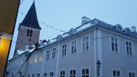 Trabajador-En-El-Elevador-Trabaja-Para-Quitar-La-Nieve-Y-El-Hielo-Del-Techo-En-El-Fondo-Podemos-Ver-La-Iglesia-De-Tartu-Jaani