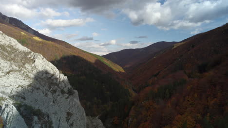 Aerial-footage-in-a-mountain-valley-with-rocks,-deciduous-and-fir-forests,-a-blue-sky-with-the-sun-and-clouds-during-the-autumn-season