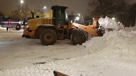 Yellow-tractor-is-removing-huge-pile-of-snow,-loading-to-red-truck-to-take-it-out-of-city