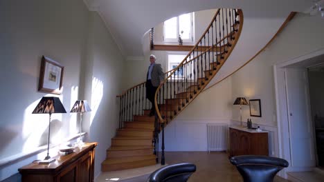 Slow-dolly-backward-shot-of-a-senior-man-coming-down-a-spiral-staircase-in-his-chateau