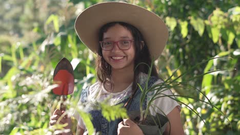 Una-Chica-Sonriente-Con-Sombrero-Y-Gafas,-Parada-En-Medio-De-Un-Jardín-Botánico-Con-Una-Pala-Y-Una-Planta-De-Cebollino-En-Las-Manos.