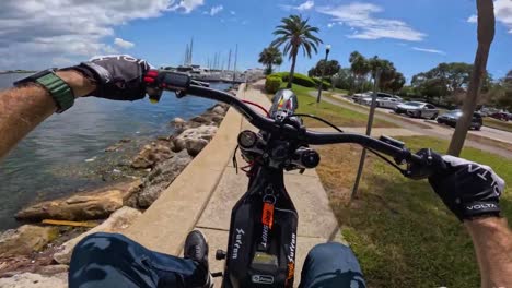 Hombre-En-Ebike-Realizando-Acrobacias-Cerca-De-Un-Cuerpo-De-Agua,-Fpv-De-Hombre-Haciendo-Caballitos-En-La-Acera