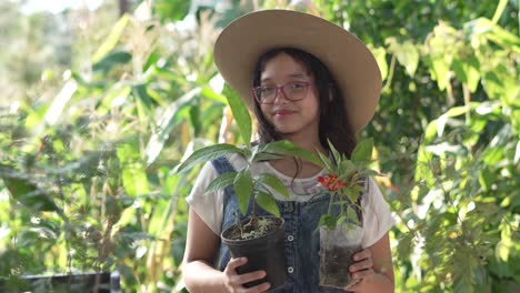 Smiling-girl-wearing-a-hat-and-glasses,-standing-in-the-middle-of-a-botanical-garden-with-an-avocado-plant-and-a-pot-of-flowers-in-her-hands
