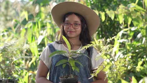 Smiling-girl-wearing-a-hat,-coverall-and-glasses,-standing-in-the-middle-of-a-botanical-garden-with-an-avocado-plant-in-her-hands