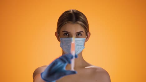 Static-shot-of-a-young-pretty-woman-or-nurse-looking-into-the-camera-and-holding-a-syringe-with-medicine-while-wearing-a-FFP2-mask-against-corona-and-other-diseases-in-front-of-orange-background