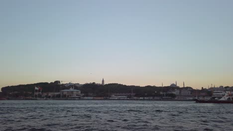 Evening,-cinematic-slow-mo,-the-view-from-a-ferry-sailing-along-the-Bosphorus-in-Istanbul,-showcasing-the-trees-and-the-iconic-backdrop-of-Hagia-Sophia-and-the-Istanbul-cityscape