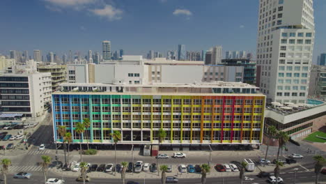 The-colorful-Dan-Hotel-in-Tel-Aviv,-Rainbow-building-on-the-Gordon-promenade-and-Frishman-beach-full-of-visitors-on-a-warm-and-calm-summer-day---pull-back-shot-to-the-sea