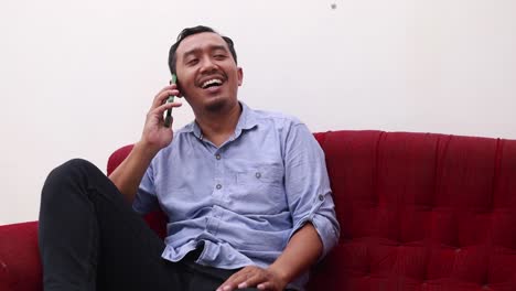 Happy-asian-adult-man-talking-on-the-phone-while-sitting-on-sofa-at-home