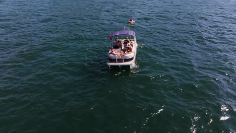 people-waving-smiling-and-having-a-good-time-on-a-pontoon-boat-pulling-an-inner-tube-on-the-lake-AERIAL-ORBIT