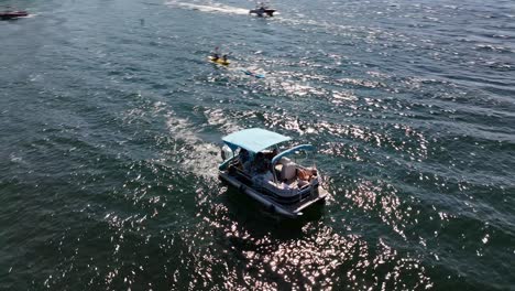 people-enjoying-their-time-relaxing-on-a-large-pontoon-boat-on-blue-water-in-lake-arrowhead-california-and-kayakers-in-the-background-AERIAL-ORBIT