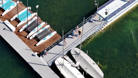 people-walking-down-a-floating-dock-crystal-clear-water-next-to-sailboats-sunny-day-AERIAL-PAN