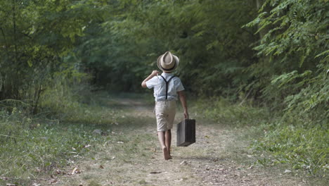 Boy-wearing-vintage-clothes-and-a-hat-walking-away-from-camera-barefoot-in-the-forest-holding-a-stick-and-a-suitcase-in-summer