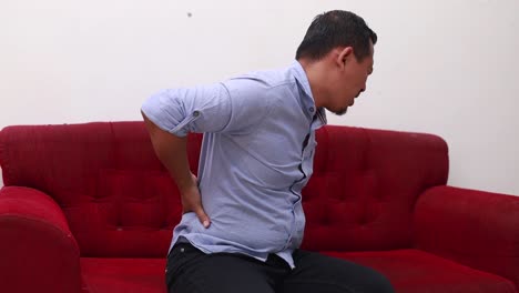 Unglücklicher-Asiatischer-Mann-Mittleren-Alters,-Der-Unter-Rückenschmerzen-Leidet,-Während-Er-Zu-Hause-Auf-Dem-Sofa-Sitzt