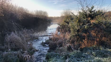 Water-flows-in-narrow-stream-on-frosty,-foggy-golden-sunrise-morning