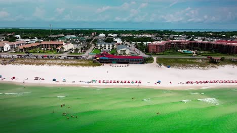 Drone-aerial-view-flying-towards-from-Pompano-Joe's-restaurant-in-Destin-Florida-with-a-view-of-old-98,-white-sand,-emerald-green-water-and-lots-of-umbrellas-and-beach-chairs-in-Destin-Florida