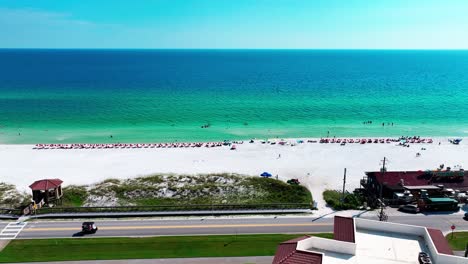Destin-Florida-aerial-shot-flying-sideways-down-old-highway-98-with-beautiful-views-of-the-Gulf-of-Mexico,-white-sand,-pompano-joes-restaurant,-golf-cart,-parking-lot,-beach-chairs,-and-umbrellas