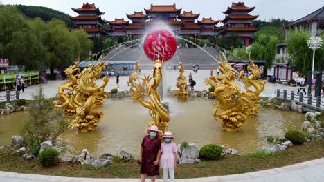 Static-slow-motion-shot-of-masked-young-girl-and-old-woman-taking-a-picture-in-front-of-water-fountain-with-gold-colored-dragons