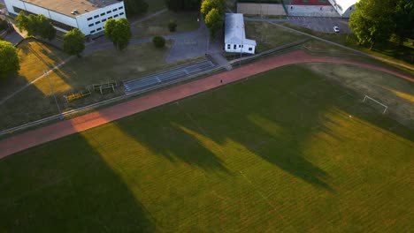 Drohnenaufnahme-Eines-Grünen-Fußballfeldes-Während-Des-Sonnenuntergangs,-Dort-Läuft-Ein-Läufer-Um-Das-Stadion-Herum