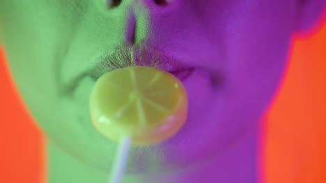 Close-up-shot-of-young-sexy-woman-lips-while-she-licks-a-sweet-lemon-lollipop-with-pleasure-with-her-tongue-against-orange-background-with-green-purple-contrast-on-face-in-slow-motion
