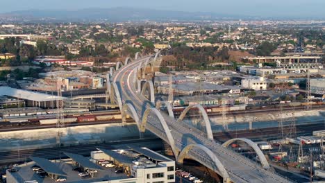 Aerial-footage-of-iconic-6th-Street-Bridge-landmark-over-the-Los-Angeles-River-on-a-clear-day,-with-cars-driving-on-a-freeway-in-the-background