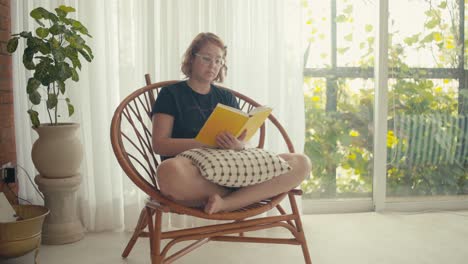 Bright-indoor-scene-with-young-woman-on-rattan-chair-reading-a-book,-medium-shot